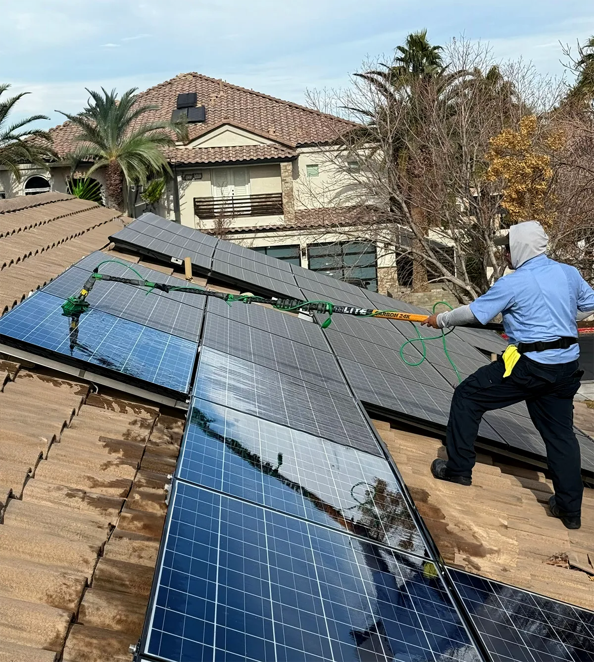A technician cleans solar panels on a roof using a long, extendable pole, with a residential home and palm trees in the background